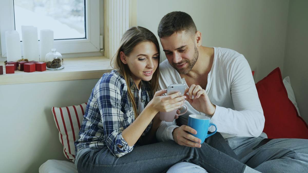 Smiling young couple talking and browsing social media on smartphone while sitting in on bed and drink coffe in the morning at home