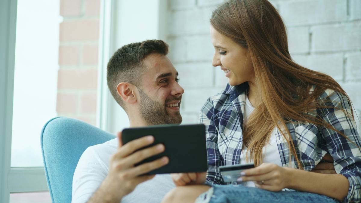 Young cheerful couple using digital tablet computer for online shopping sitting on balcony in modern loft apartment